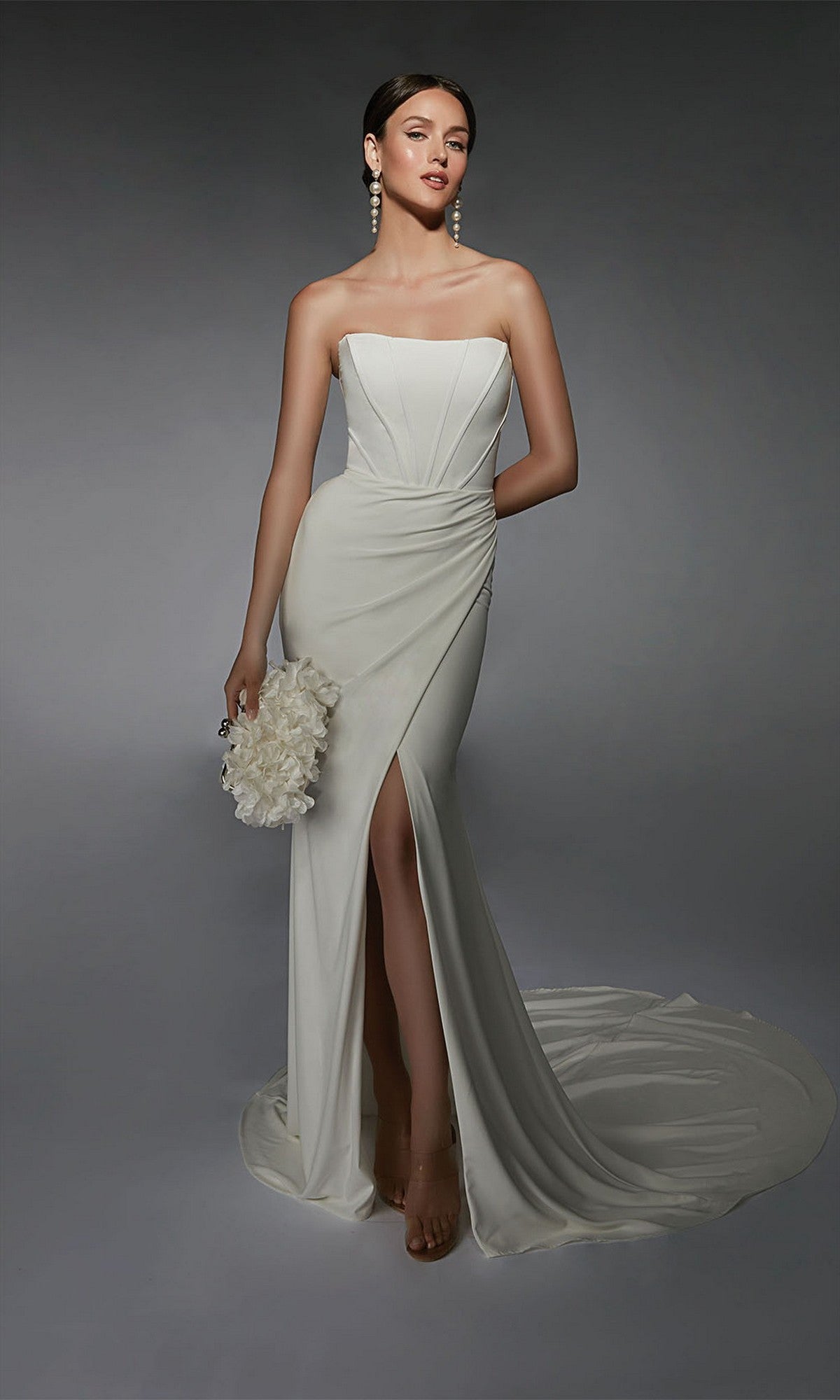 A woman wearing the Long White Dress: Alyce 7102, a strapless white bridal gown with a thigh-high slit and corset bodice, holds a bouquet of white flowers against a plain gray background.